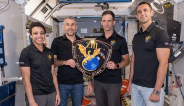 three men and one woman, all wearing black polo shirts, hold a plaque inside a mockup space module