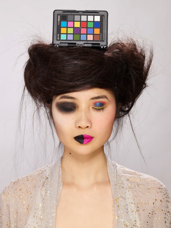 A person with voluminous dark hair balances a color checker card on their head, featuring split-style makeup with black eyeshadow and lipstick on the left and colorful, vibrant makeup on the right.