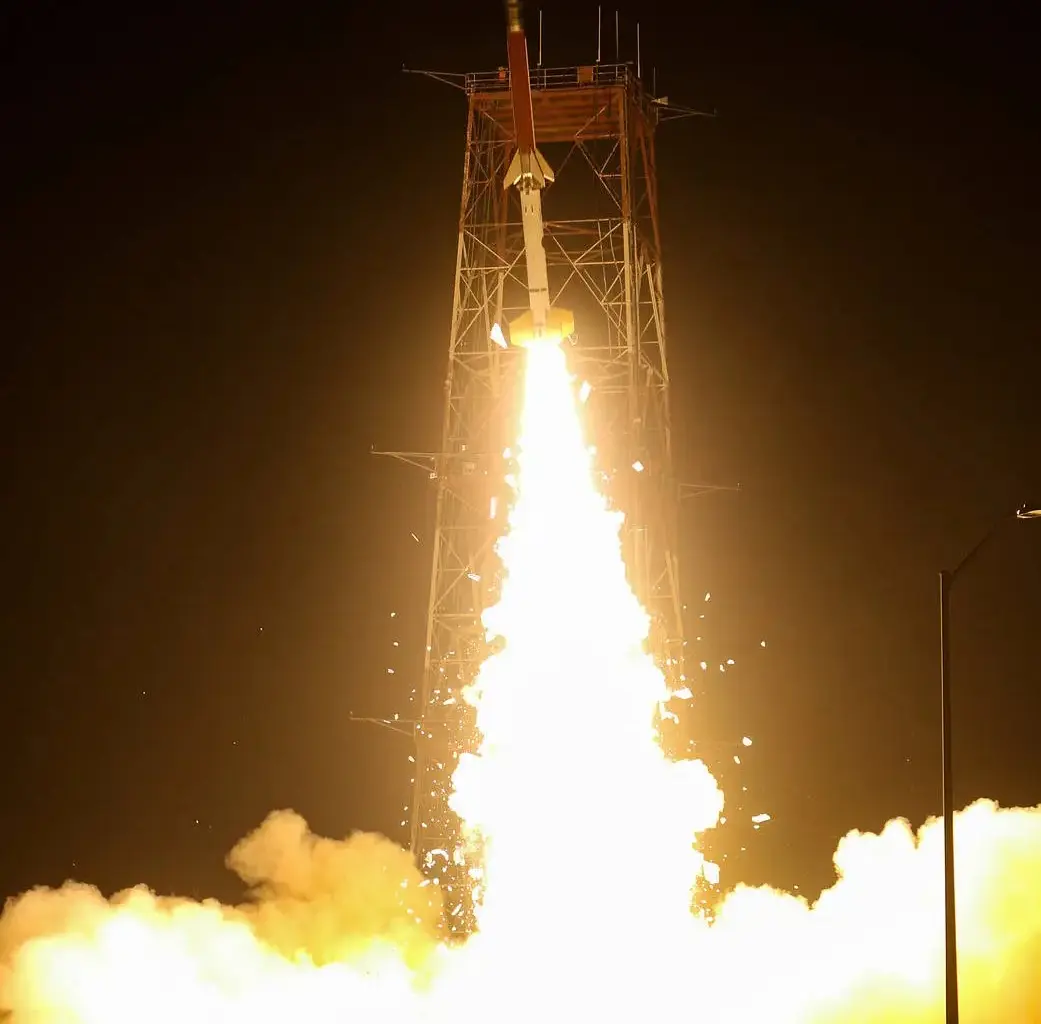 A rocket launches at night, surrounded by bright flames and smoke, with a tall supporting structure visible and the dark sky in the background.