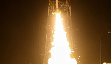 A rocket launches at night, surrounded by bright flames and smoke, with a tall supporting structure visible and the dark sky in the background.