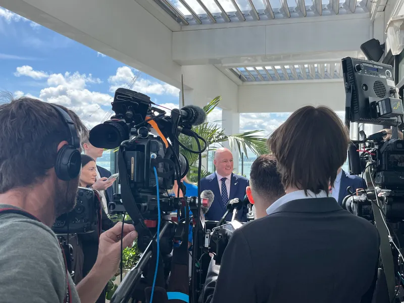 A man in a suit stands in front of TV cameras and reporters