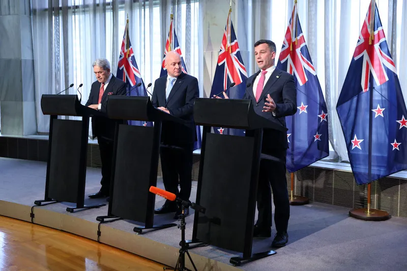 Three men in suits stand at lecterns in front of New Zealand flags