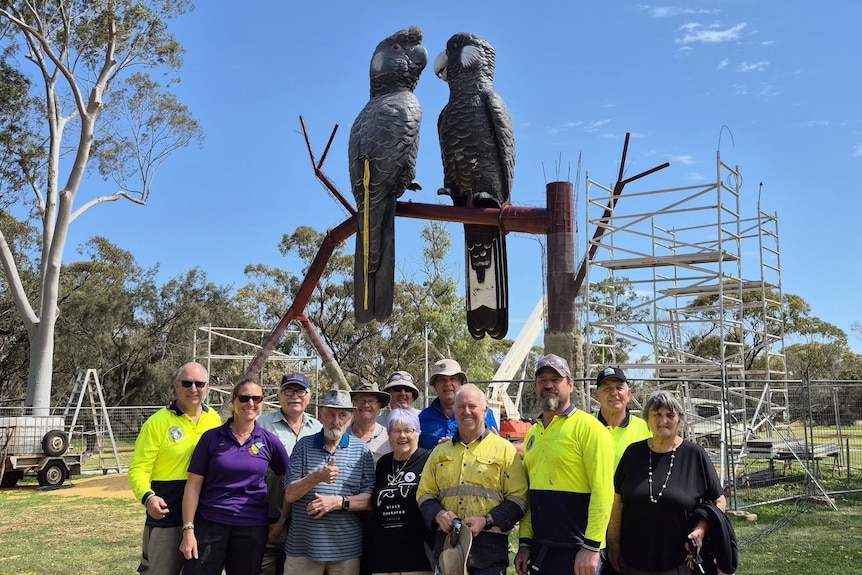 A group of people smile in front of a large sculpture of two black cockatoos perched on a branch.