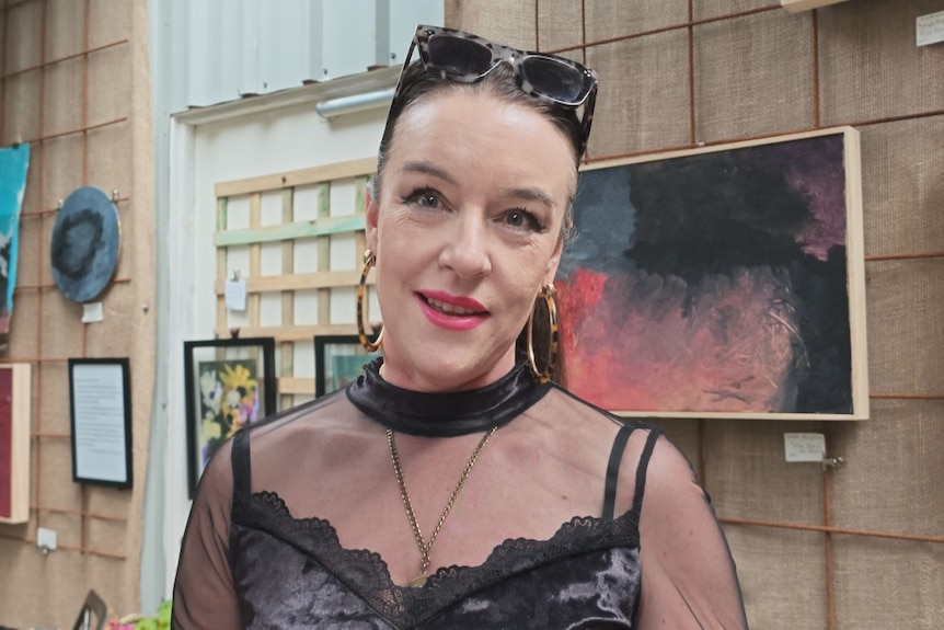 Serena McLaughlan stands with artwork on the wall behind her. She wears black, lacy clothes and large gold hoop earrings.