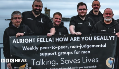 A group of men in black tops standing on Douglas Promenade holding a banner about the support services that says talking saves lives.