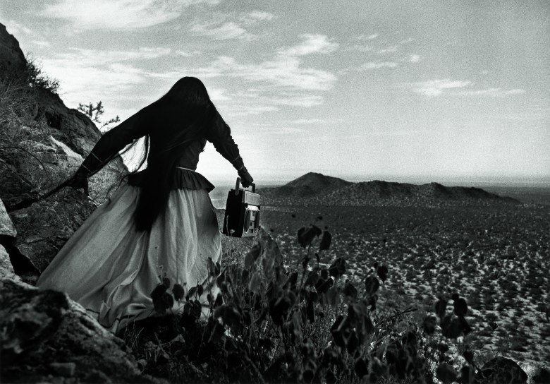 A person in a long skirt stands on rocky terrain holding a boom box, looking out over a vast desert landscape with hills in the distance.
