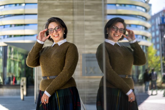 Monfared smiles and adjusts her glasses with one hand outside a curved glass building, her reflection visible in a window beside her.