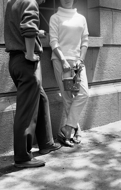 Two people stand on a sidewalk by a building. One holds a small handbag and wears heeled sandals, cropped pants, and a sweater; the other wears dark pants and loafers. Their faces are not visible.