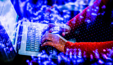 Hands typing on a laptop keyboard, overlaid with a double exposure of glowing blue digital light and data patterns.