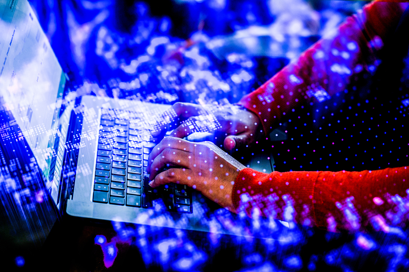Hands typing on a laptop keyboard, overlaid with a double exposure of glowing blue digital light and data patterns.