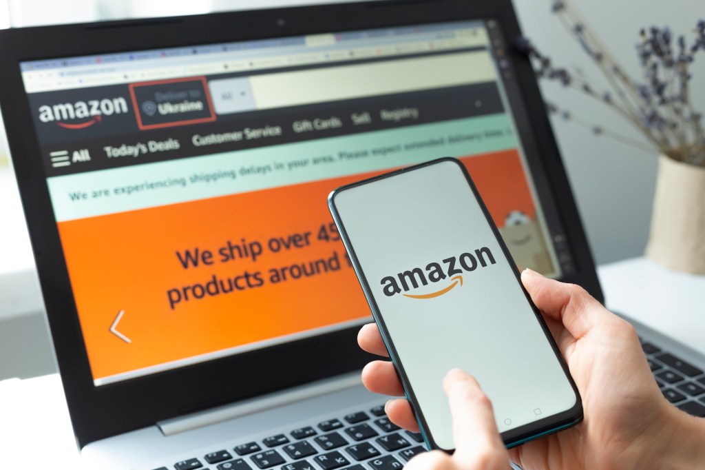 A person using a smartphone with the Amazon app open, with a laptop displaying the Amazon website in the background.