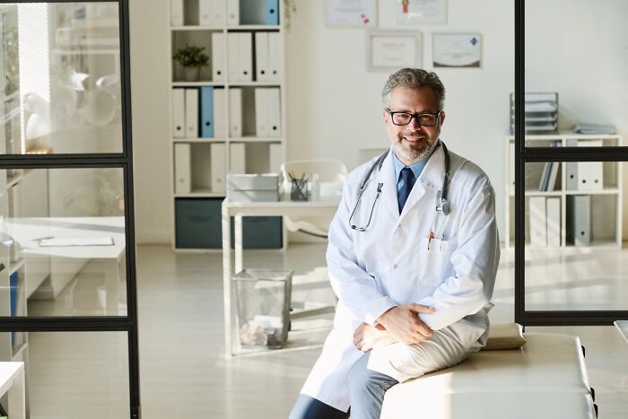 A smiling doctor in glasses sitting on an examination table in a bright medical office.