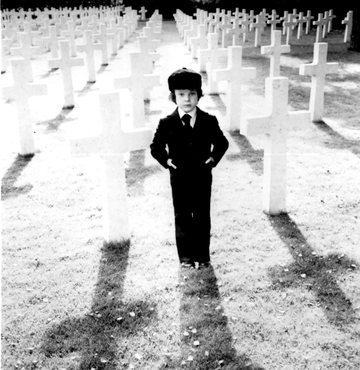 A young boy in suit, both hands in pockets, stands in a cemetery