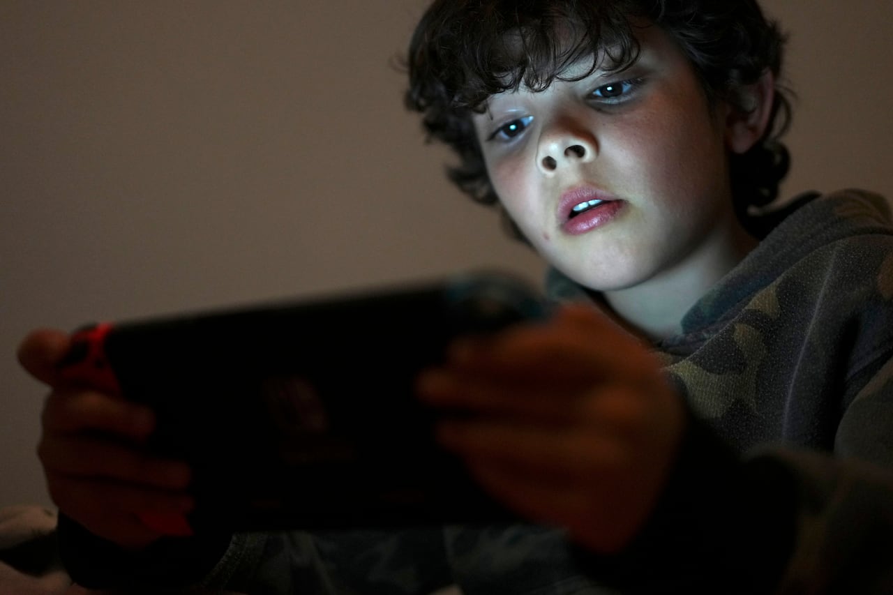 A young boy with brown, curly hair looks at the lit screen of a smartphone that he's holding in his hands. The glow of the screen is reflected on his face in a darkly lit room.