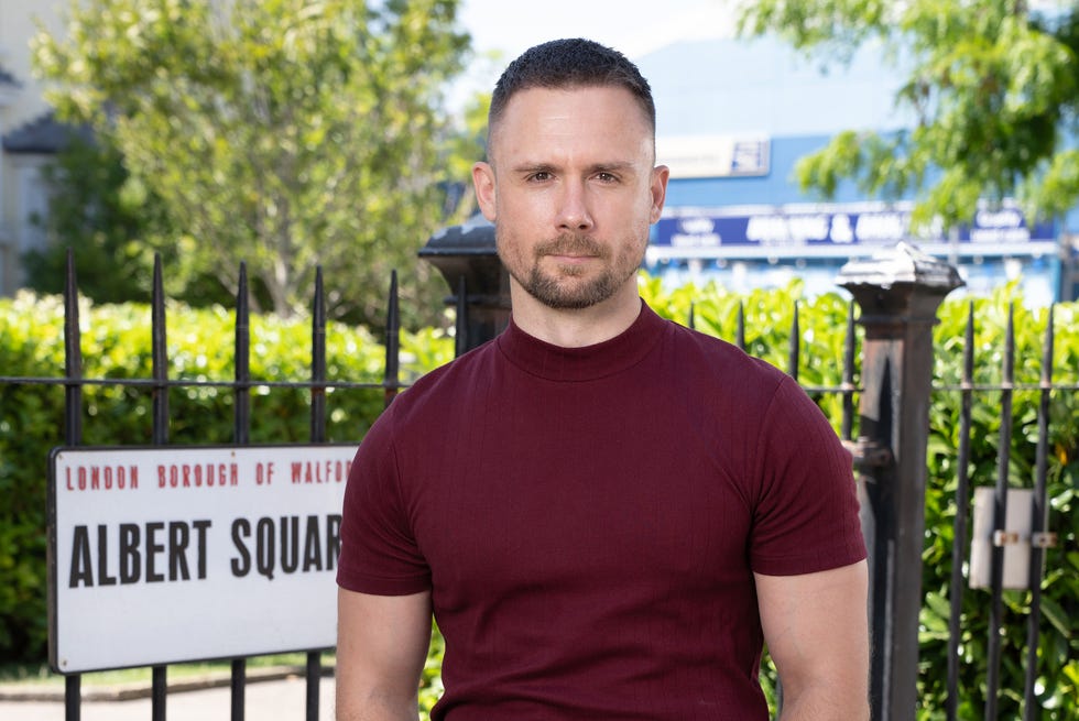 EastEnders, Lee Carter in 2026 eastenders, danny hatchard as lee carter in 2026, wearing a red tshirt standing outside the square gardens
