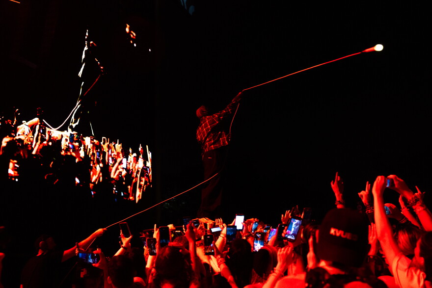A man in a mask swings a microphone on a long cord over a crowd at night. Everyone in the image is awash in red light.
