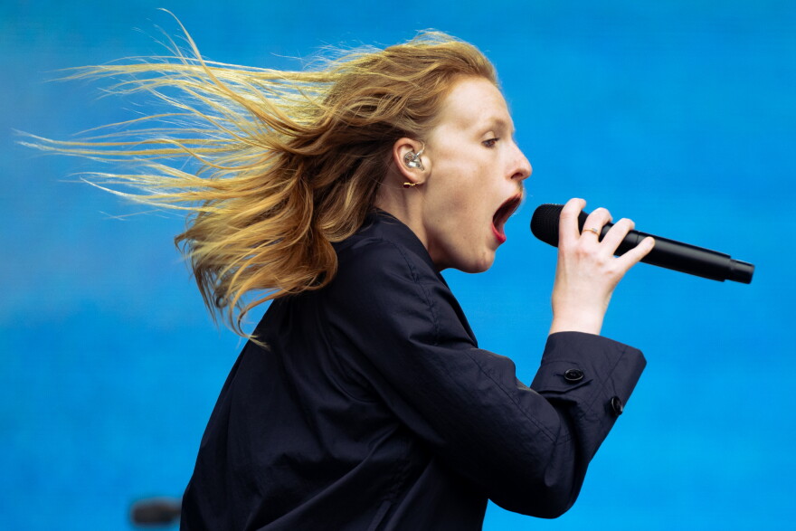 A woman is seen from the side singing into a microphone as her hair is straight out behind her. In the background is a blue screen.