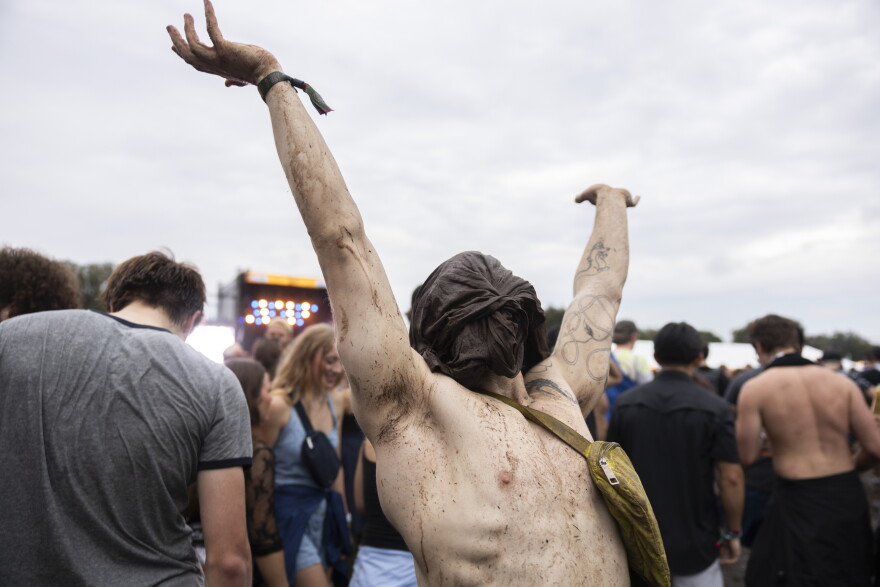 A shirtless man wearing a shirt tied around his face, while covered in mud, raises his arms into the air in a crowd.