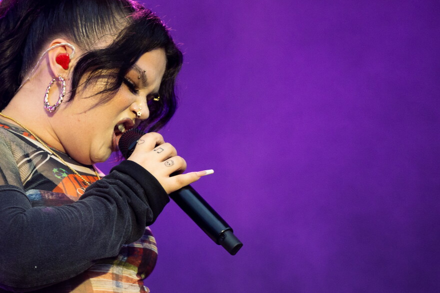 In front of a purple background a woman, shown closely, snears into a microphone.