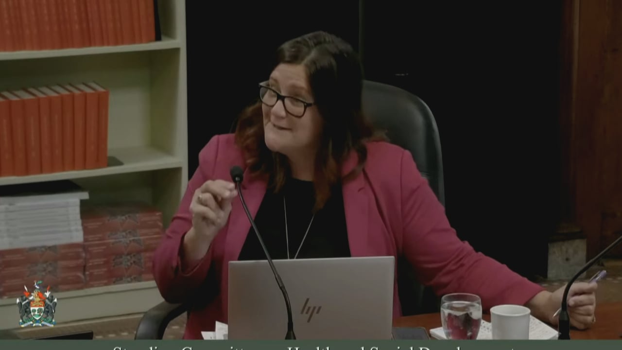 A woman in glasses and a pink coat gestures while sitting in front of a laptop computer.