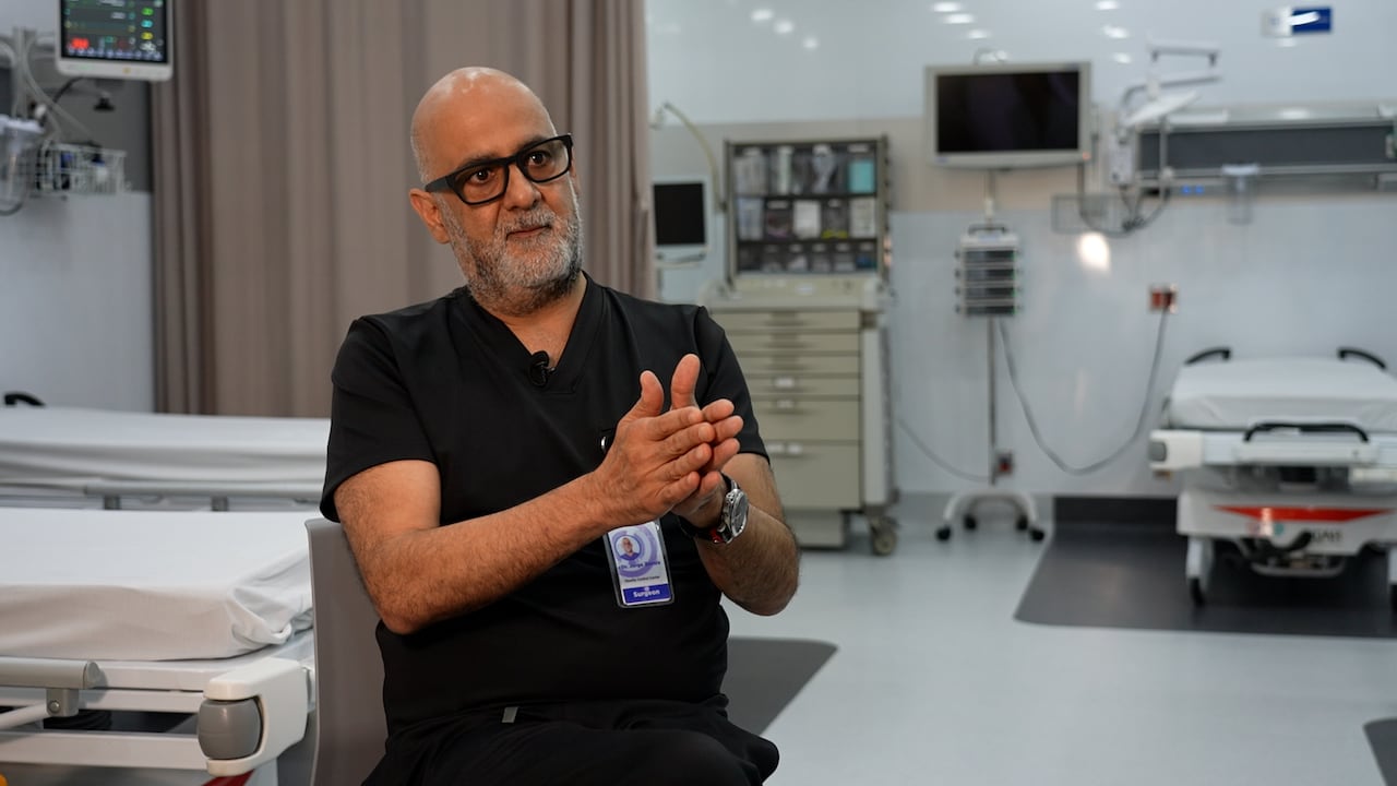 A man in doctor scrubs sits on a chair and gestures with his hands.