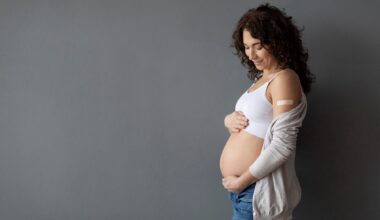 Happy Pregnant Female With Adhesive Bandage On Shoulder Embracing Her Belly While Standing Over Grey Studio background,