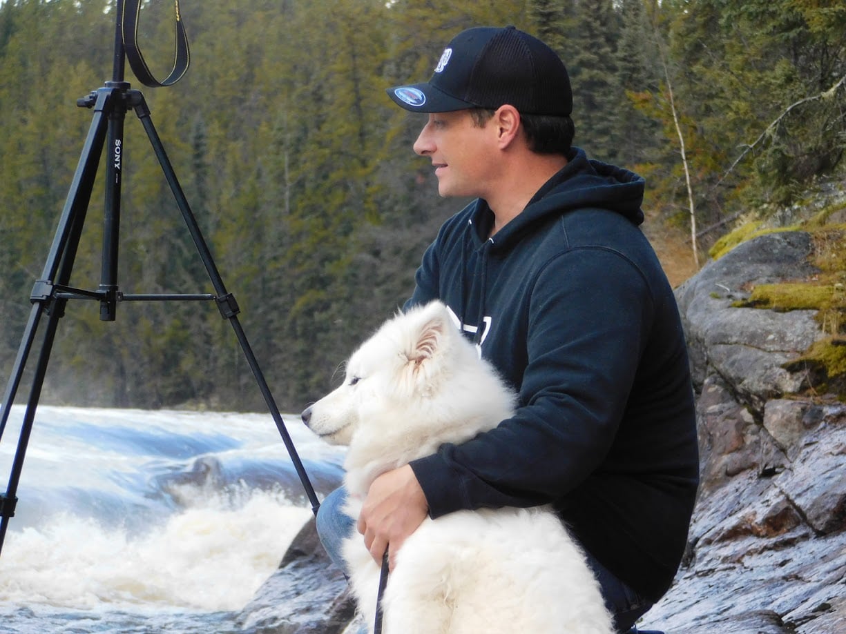 A man and a white dog sit by a river with a tripod.