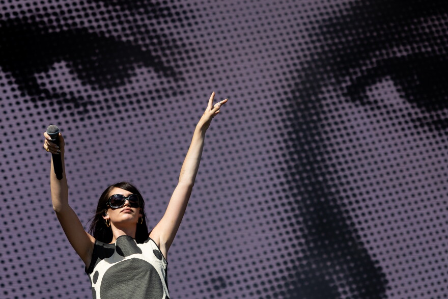 A woman raises her hands in the air as she performs on a stage with a display of a face behind her.