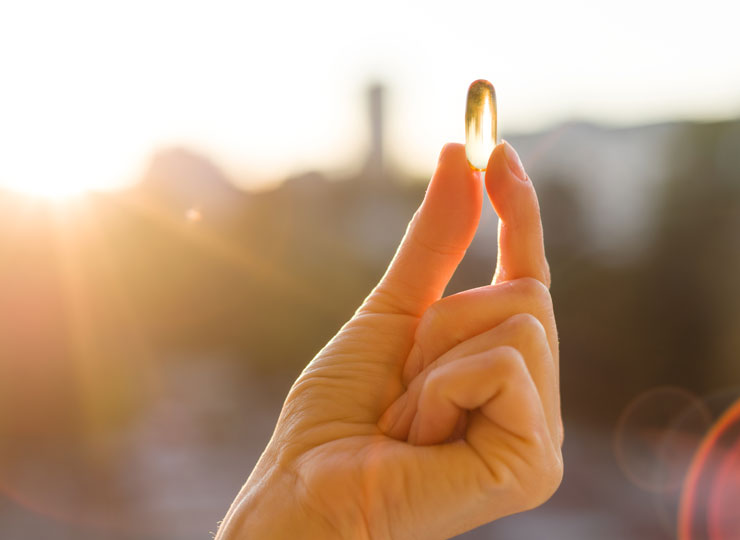 A hand holding a vitamin D softgel up to the sunlight