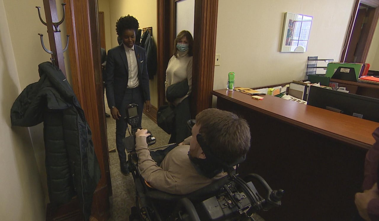 A man in a beige sweater, seated in a wheelchair, meets a person with a blue suit and buttoned-up grey shirt.