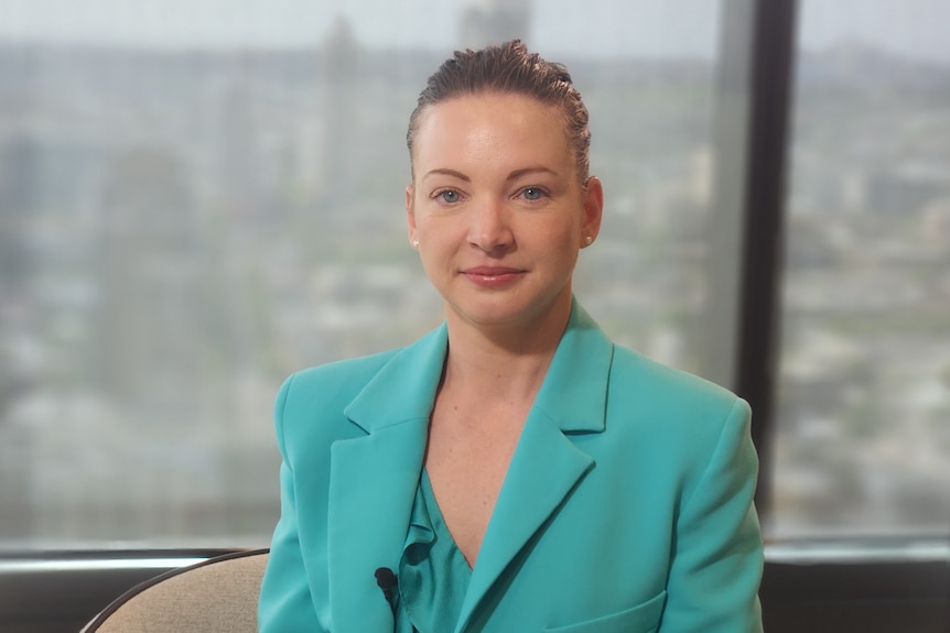 Nicola Powell, wearing a blue-green suit, sits in a chair in front of a window.