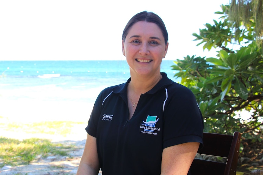 A young woman smiles broadly while sitting on a bench with the ocean behind. 