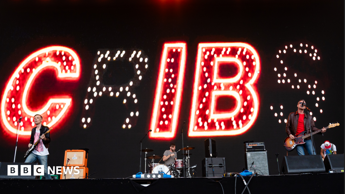 The band The Cribs play on a big stage. Lights behind spell the band name.