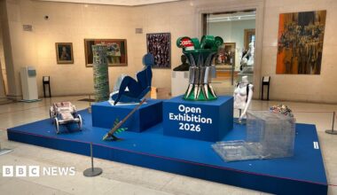 An exhibit in a gallery showcases a blue platform in the middle with several quirky items of art placed on it. On the walls surrounding it are several paintings of different sizes on the walls. The display reads Open Exhibition 2026 in the middle.