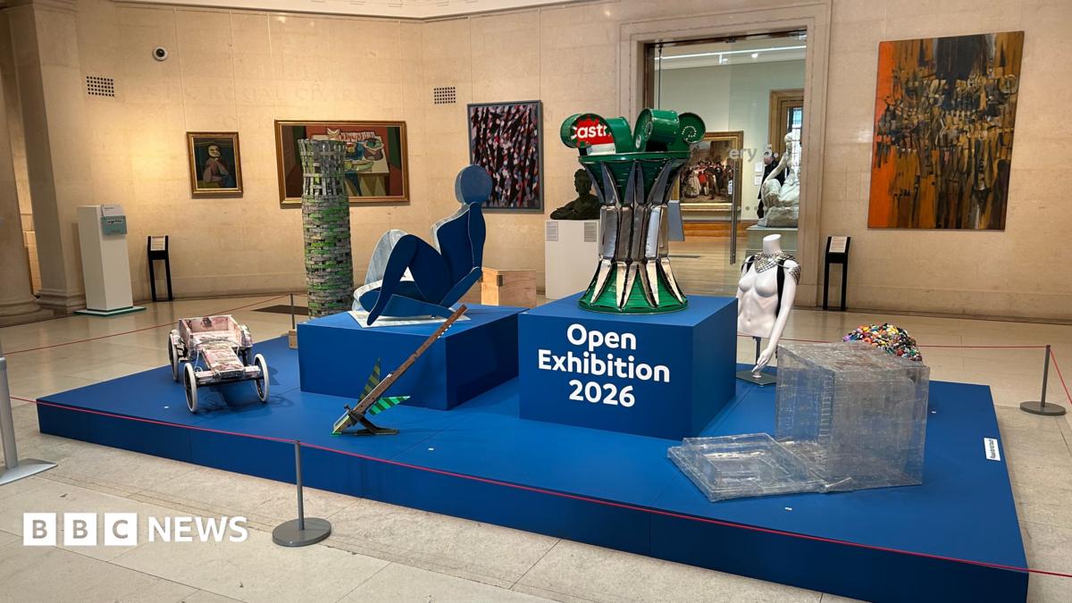 An exhibit in a gallery showcases a blue platform in the middle with several quirky items of art placed on it. On the walls surrounding it are several paintings of different sizes on the walls. The display reads Open Exhibition 2026 in the middle.