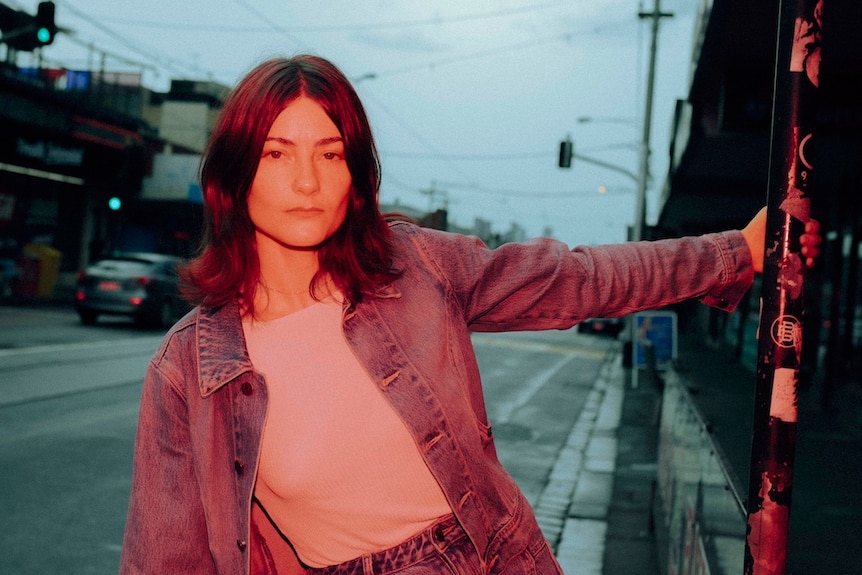 A dark-haired woman in denim jacket, jeans and a white top, leans off a pole in street.