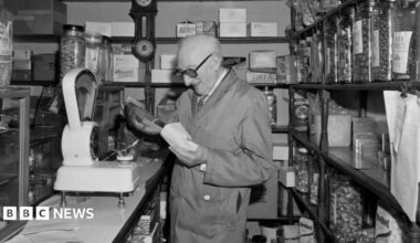 A black-and-white photograph of a traditional sweet shop interior. A shopkeeper wearing a work coat stands behind the counter reading a paper bag, beside a large mechanical weighing scale. Shelves lining both sides of the narrow shop are filled with jars of sweets and confectionery boxes, while a wall-mounted clock hangs behind the counter.