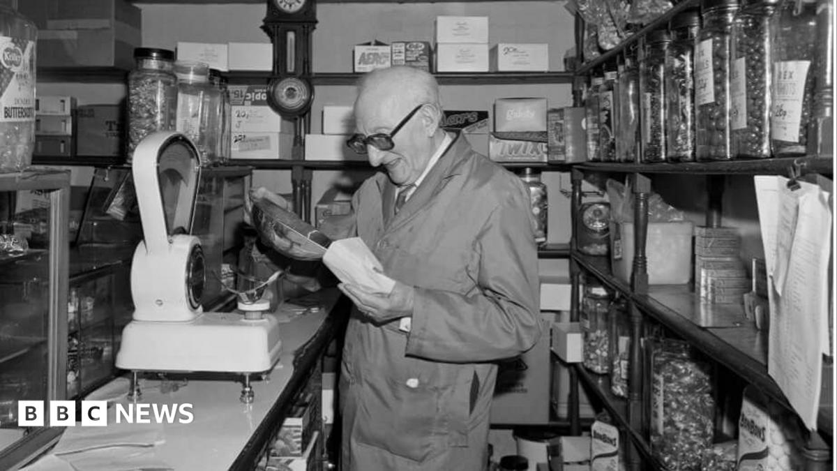 A black-and-white photograph of a traditional sweet shop interior. A shopkeeper wearing a work coat stands behind the counter reading a paper bag, beside a large mechanical weighing scale. Shelves lining both sides of the narrow shop are filled with jars of sweets and confectionery boxes, while a wall-mounted clock hangs behind the counter.