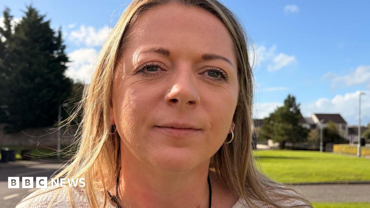 Close-up portrait of a woman with a nose ring and hoop earrings standing outdoors on a sunny day, with a grassy area, trees and houses blurred in the background.