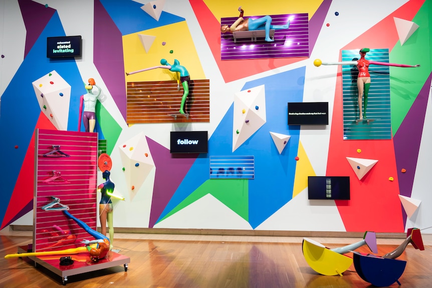 A group of mannequins, painted brightly and manipulated into strange shapes, are attached to yoga mats on a gallery wall.