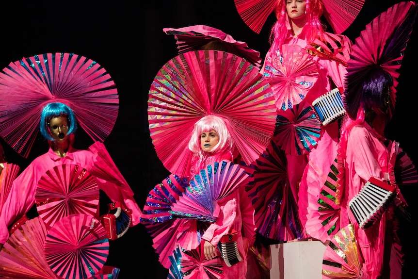 On stage, a woman in a pink dress and fanned headdress and pink bob wig, stands next to a mannequin in the same outfit.