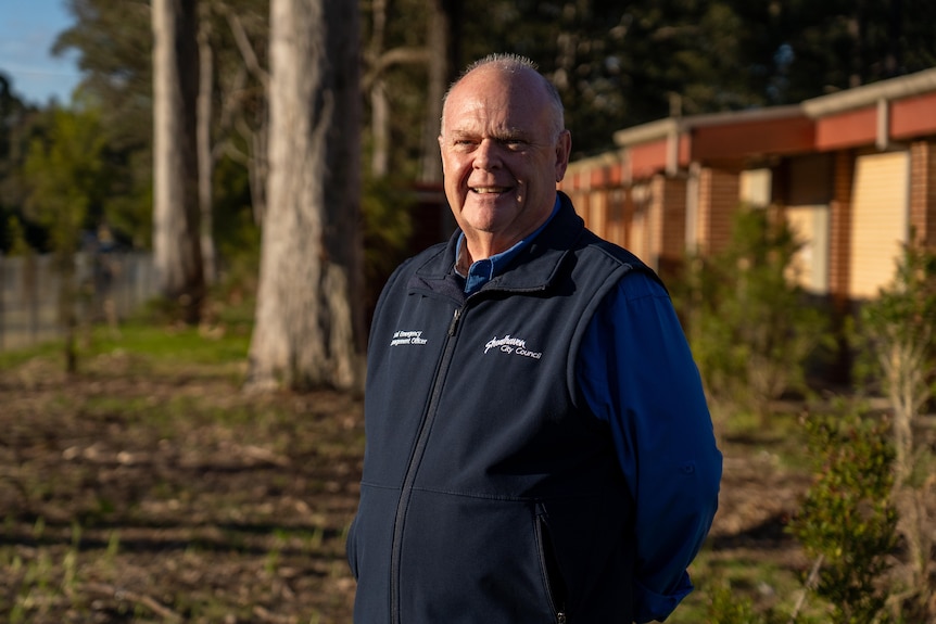 A man in a blue shirt with a blue fleece vest stands in front of trees and a red brick building