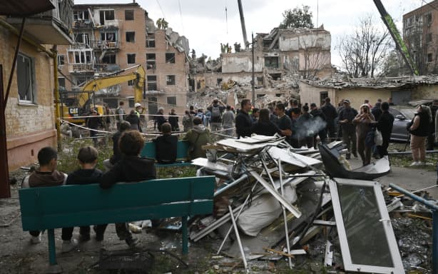 Local residents look at rescuers working on a multi-floor residential building destroyed following a Russian aerial strike in Pryluky on October 7, 2026, amid the Russian invasion of Ukraine. In Pryluky, the death toll has risen to nine people, including three children, and 40 people are known to have been injured, including seven children, the Emergency Service said on October 7, 2026. (Photo by Genya Savilov / AFP)