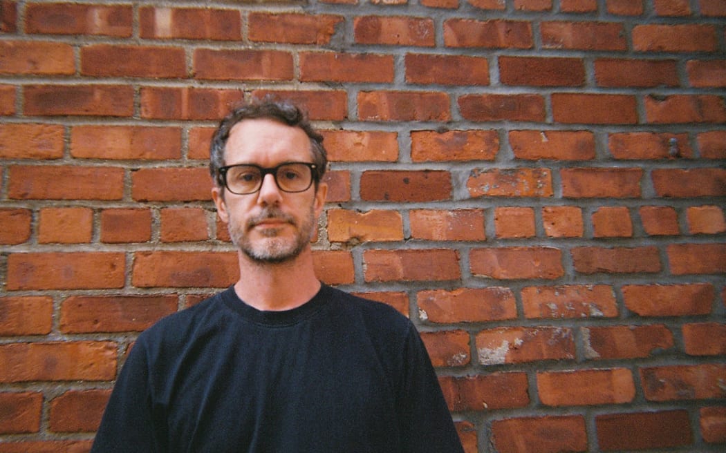 Tony Stamp is a slightly smiling man with glasses and a beard, wearing a black sweater and standing against a brown brick wall.