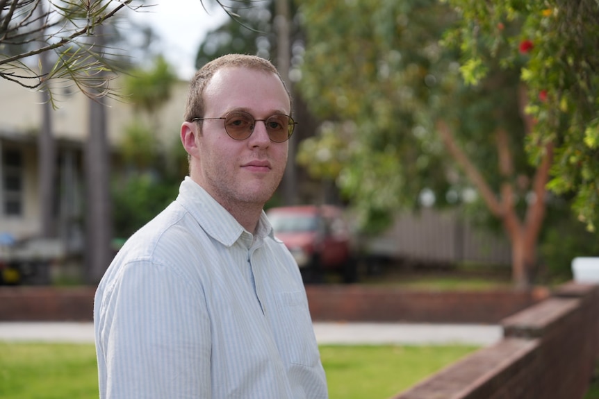 Jonathon leans on a brick wall while posing for a photo. He has short hair and dark round glasses.