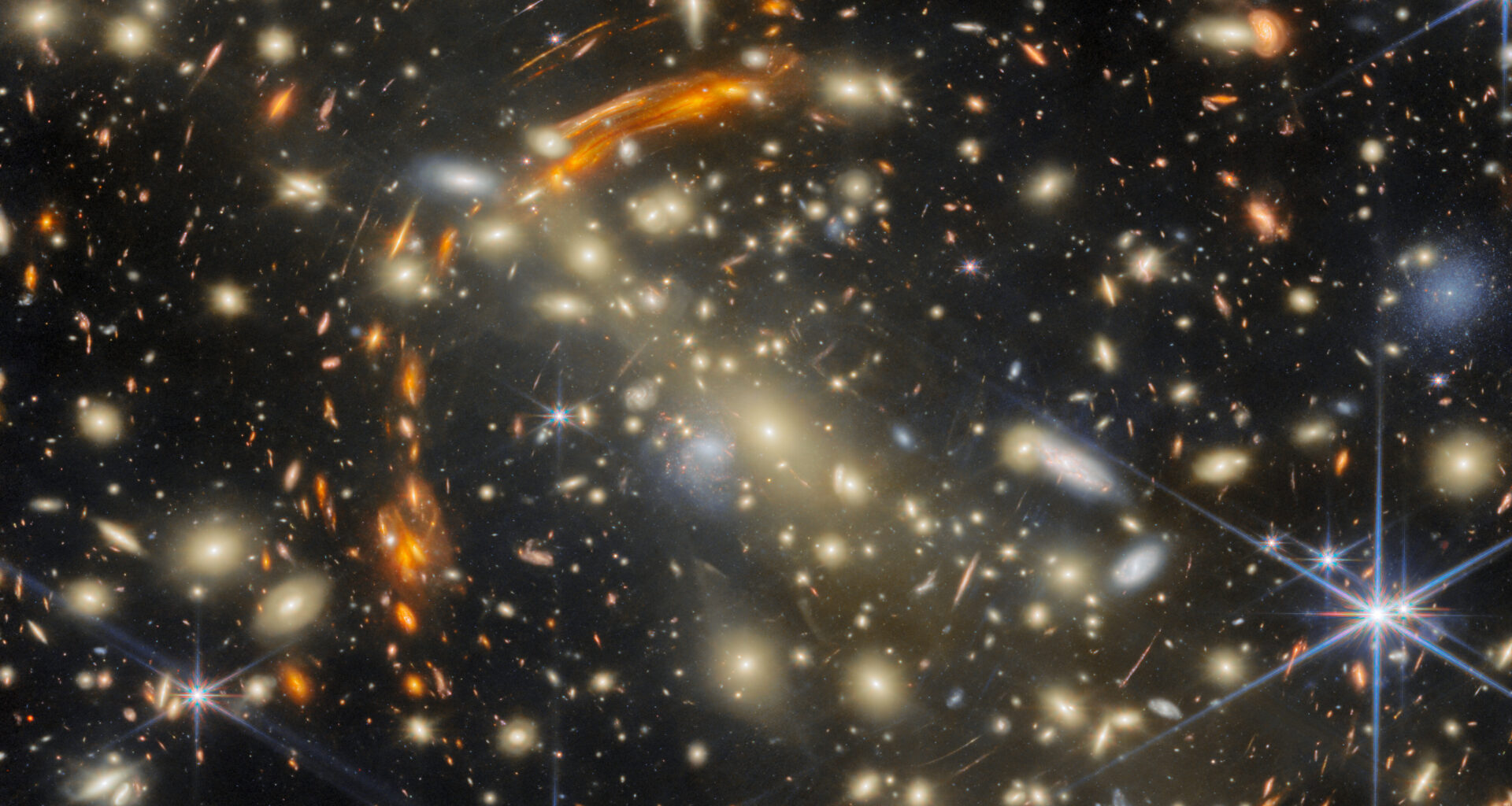 A massive galaxy cluster, made of many golden elliptical galaxies with the largest, brightest one in the center. Heavily distorted orange galaxies appear all around the cluster, the largest concentrated in a large arc around the cluster's center; these are images of very distant galaxies, created by gravitational lensing. A few galaxies are large and bluish-white in color; bright, nearby stars are the same color.