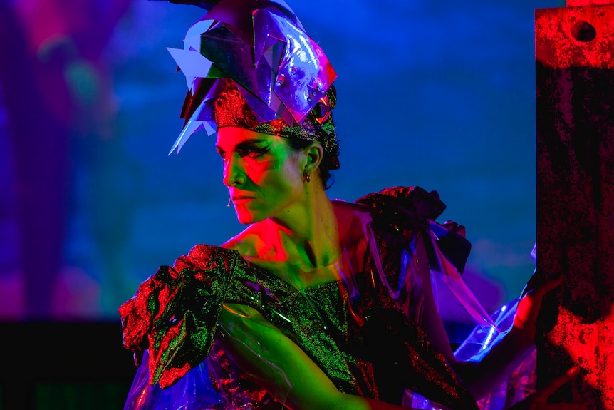 A person with a severe expression, looking to the side, wearing an ostentatious headdress and matching dress of futuristic blue.