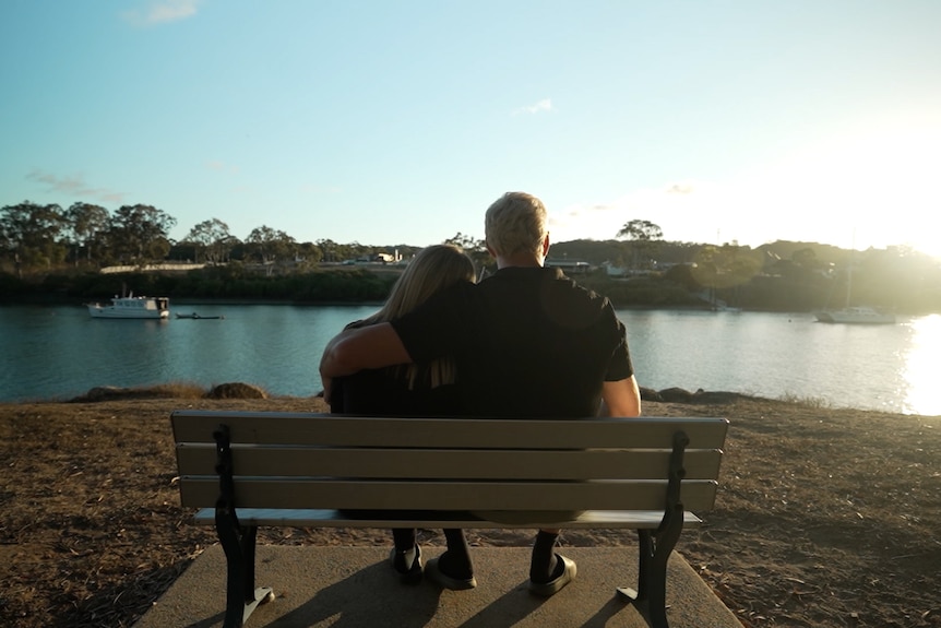 A shot from behind a bench in front of a river as two people sit shortly before sunset. The man has his arm around the woman.