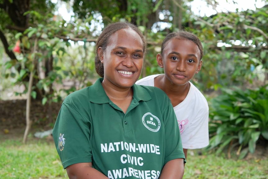 Anna Bugotu, wearing a green shirt, smiles. Her daughter leans from behind her, peering at the camera.