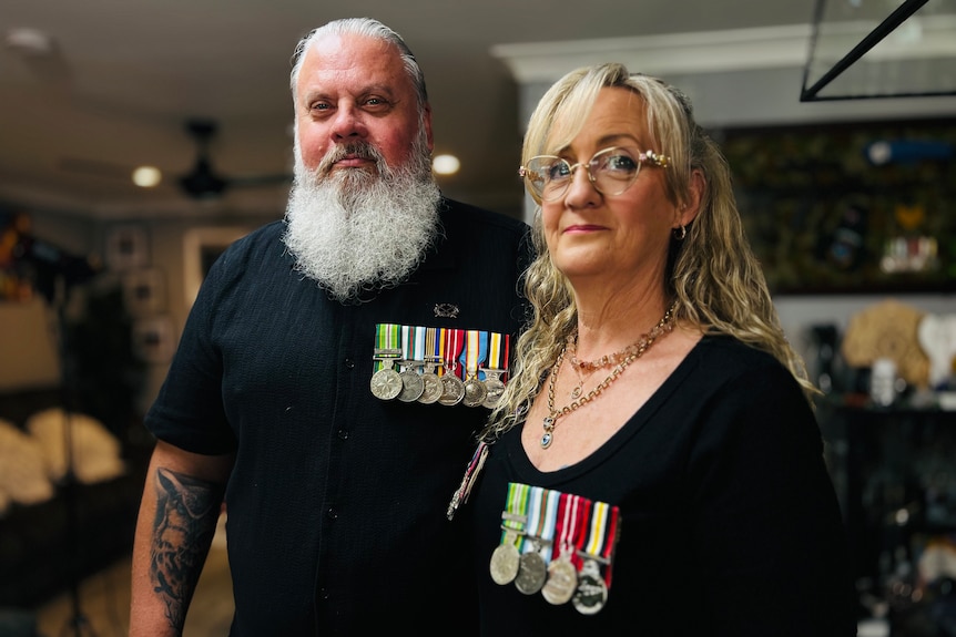 Two people stand inside a home wearing black but with several military medals on their chests. They look at the camera.